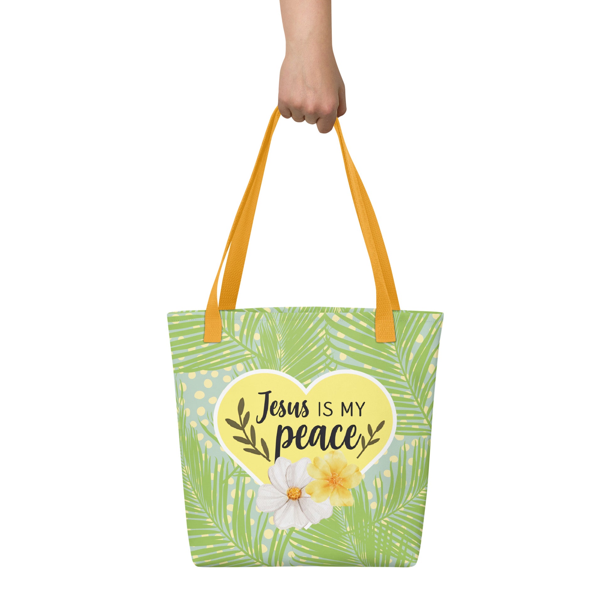Handheld view of the tote bag with yellow handles and tropical palm-leaf pattern, featuring a yellow heart that reads “Jesus is my peace” with flowers.