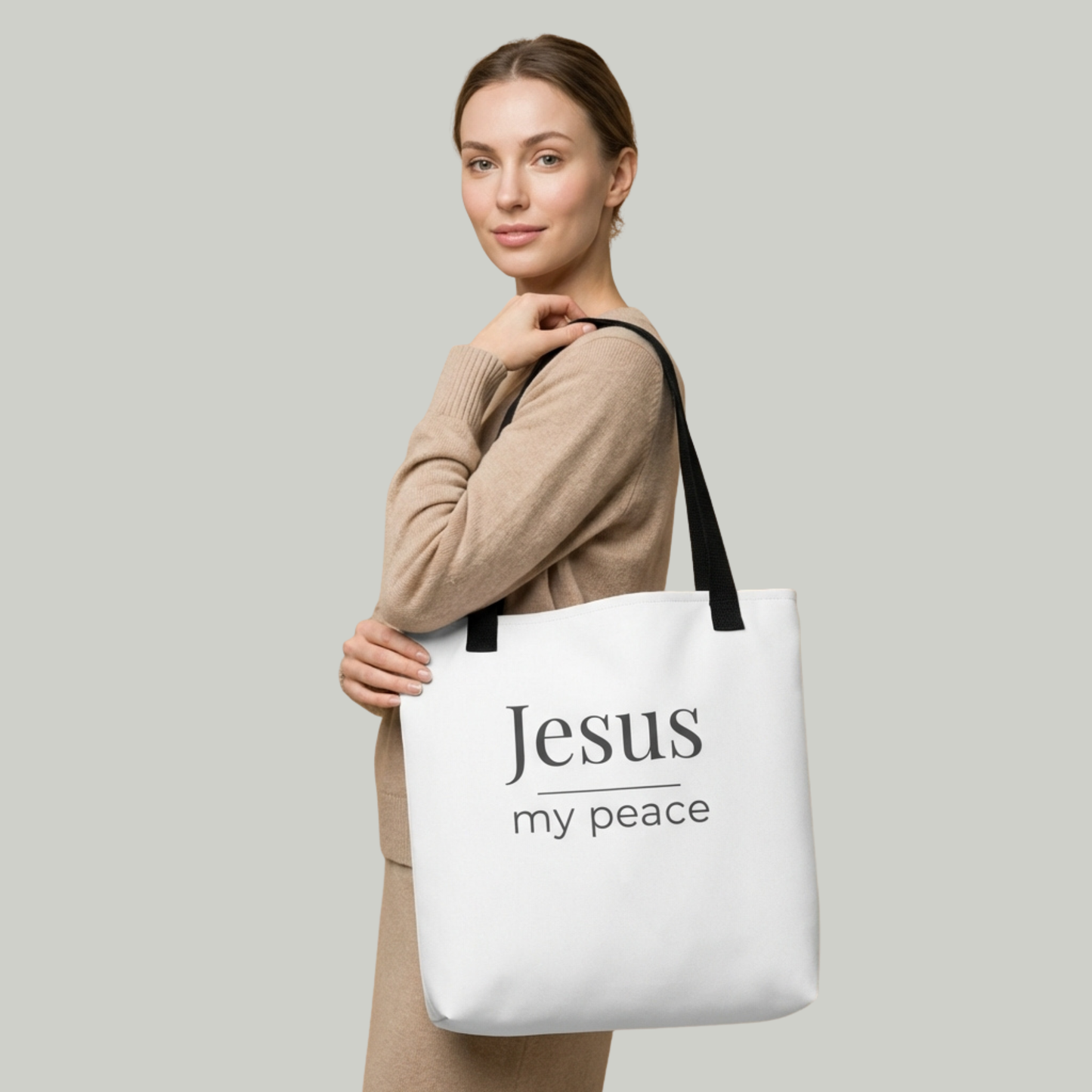 Lifestyle photo of a model holding a white tote bag with black handles featuring the text “Jesus my peace” in dark gray.