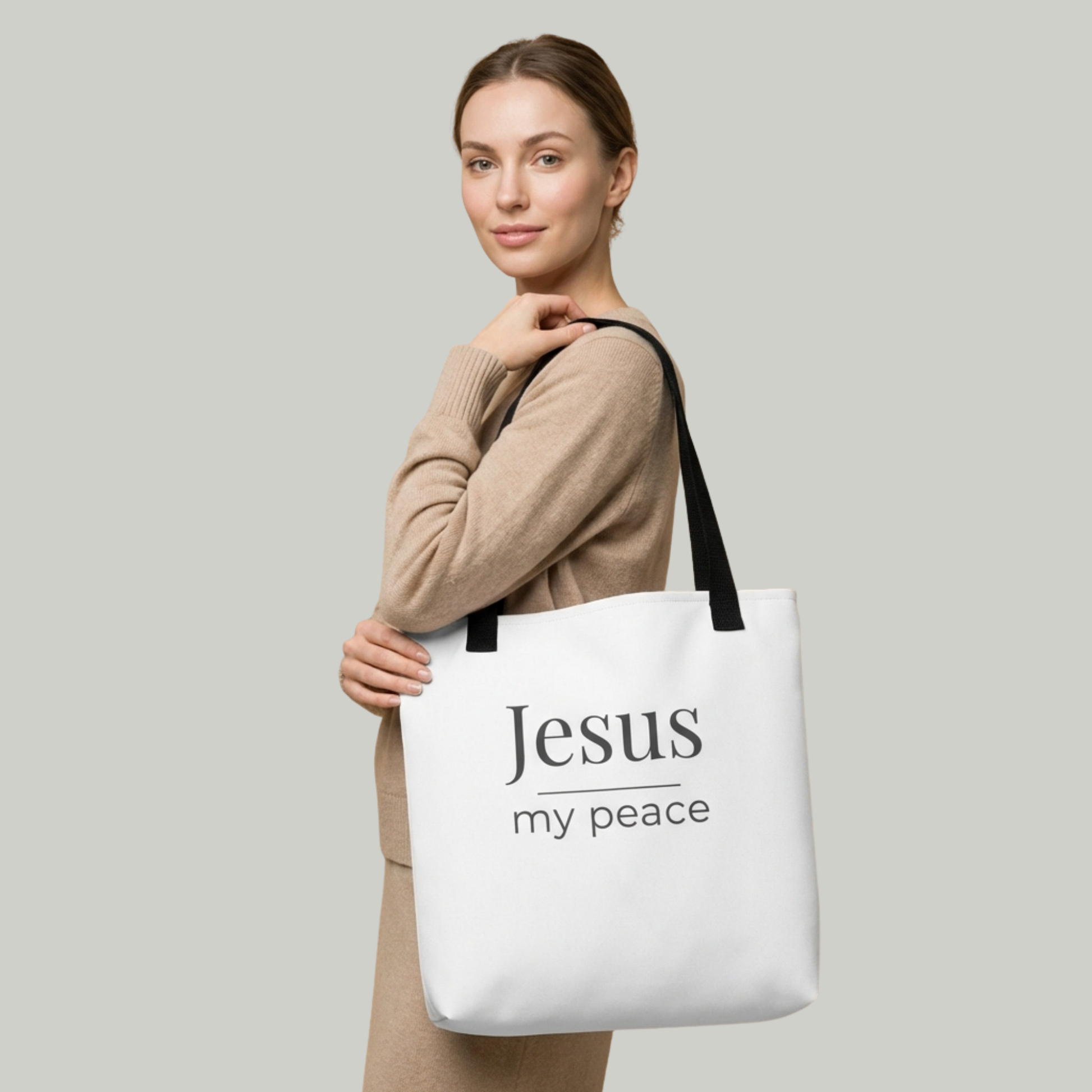 Lifestyle photo of a model holding a white tote bag with black handles featuring the text “Jesus my peace” in dark gray.