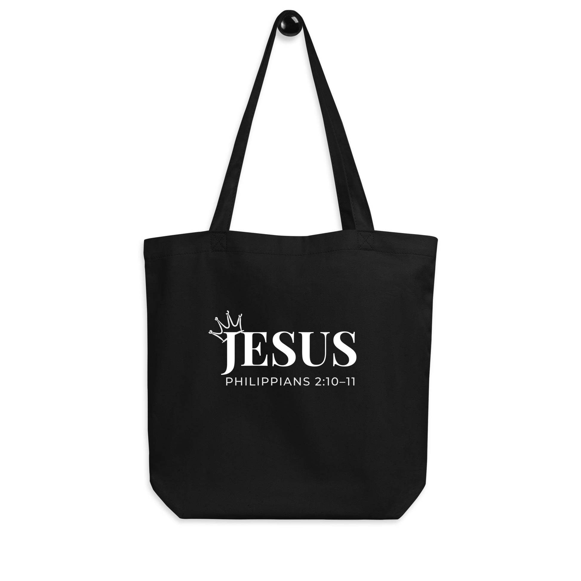 Black cotton tote bag hanging on a hook featuring white “JESUS” text with a crown detail and “Philippians 2:10–11.”