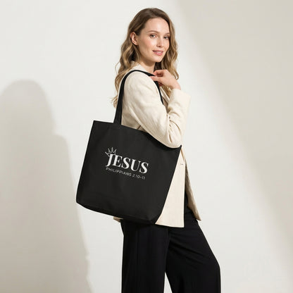 Model holding a black cotton tote bag printed with “JESUS,” a crown detail, and “Philippians 2:10–11” in white text.