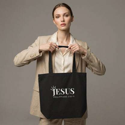 Model holding a black cotton tote bag printed with “JESUS,” a crown detail, and “Philippians 2:10–11” in white text.