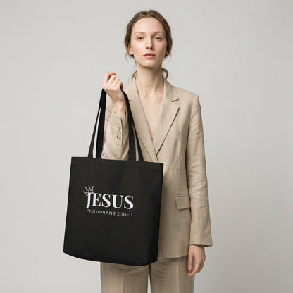Model holding a black cotton tote bag printed with “JESUS,” a crown detail, and “Philippians 2:10–11” in white text.