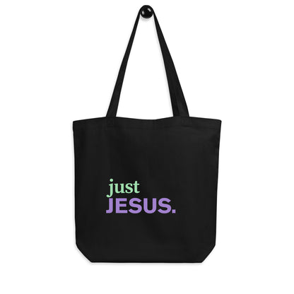 Black cotton tote bag hanging on a hook featuring the “just JESUS.” design in green and purple lettering.