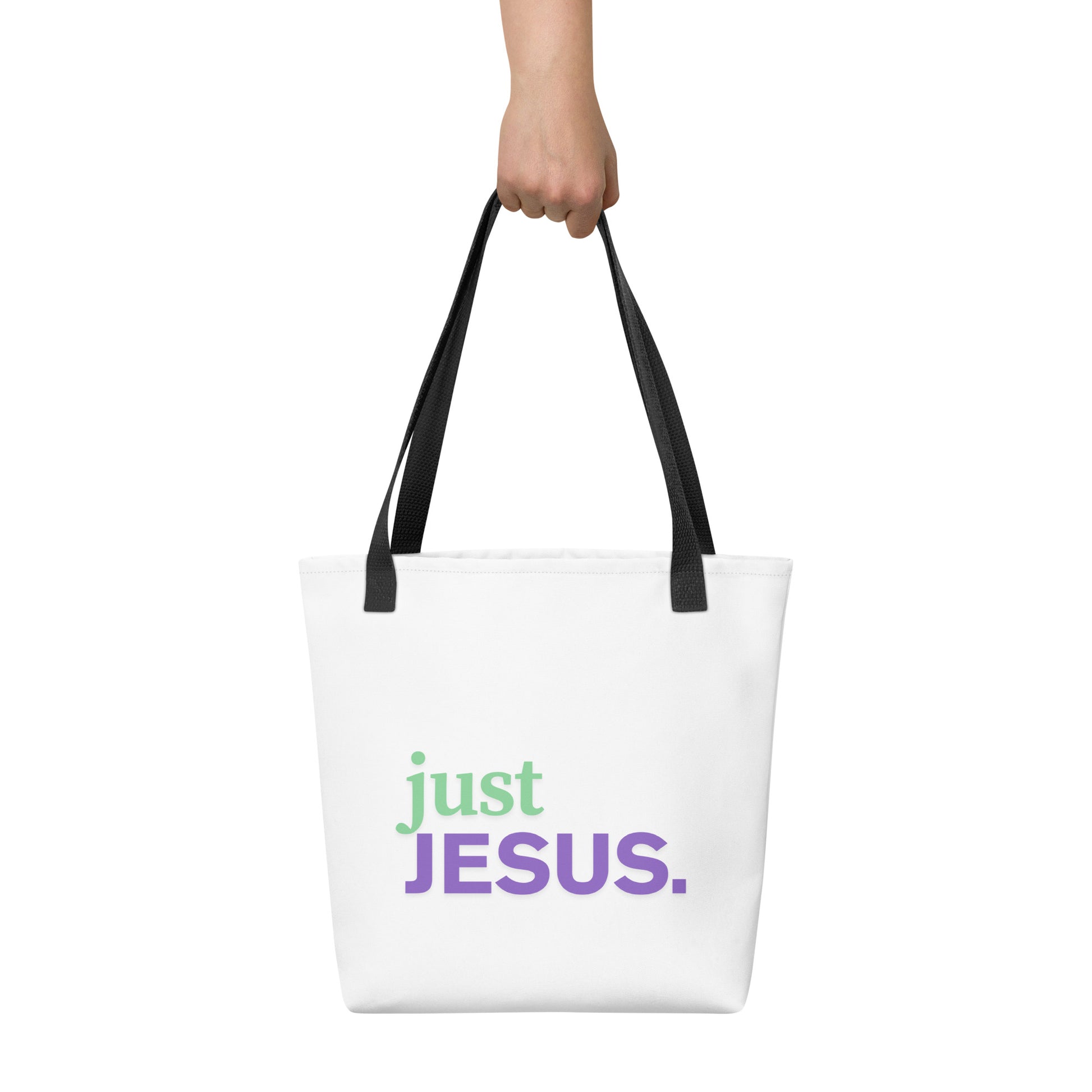 White tote bag held in hand with black straps featuring “just JESUS.” in green and purple text.