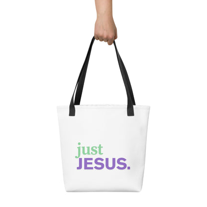 White tote bag held in hand with black straps featuring “just JESUS.” in green and purple text.