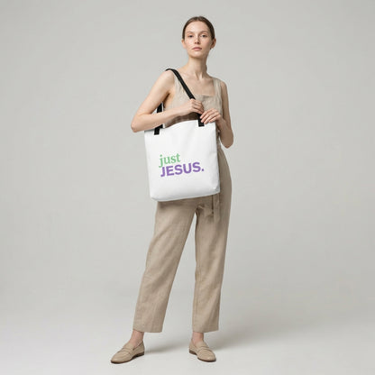 Model holding a white tote bag with black handles that reads “just JESUS.” in green and purple.