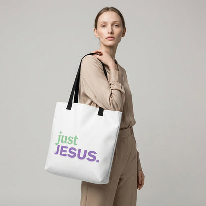 Model holding a white tote bag with black handles that reads “just JESUS.” in green and purple.