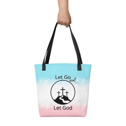 Tote bag held in hand with black handles, a pastel blue-to-pink gradient, and “Let Go and Let God” with a mountain and cross graphic.