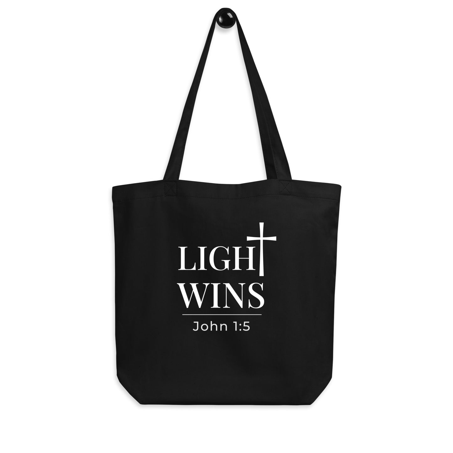 Black tote bag hanging on a hook featuring “Light Wins,” a cross graphic, and “John 1:5” in white text.