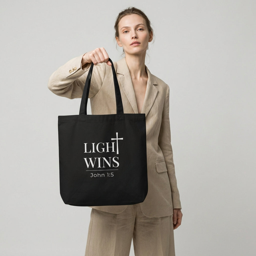 Lifestyle photo of a model holding a black tote bag featuring “Light Wins,” a cross graphic, and “John 1:5.”