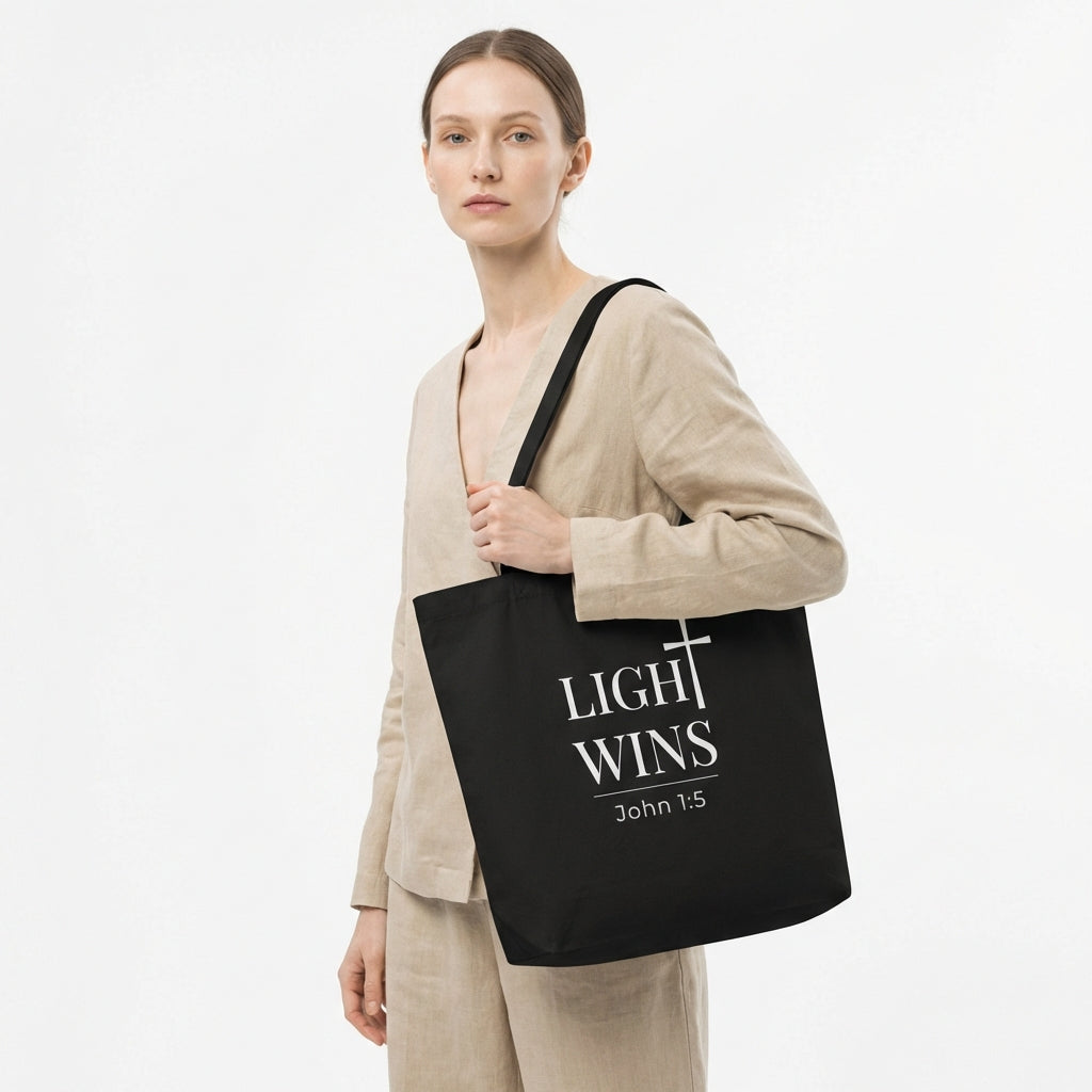 Lifestyle photo of a model holding a black tote bag featuring “Light Wins,” a cross graphic, and “John 1:5.”