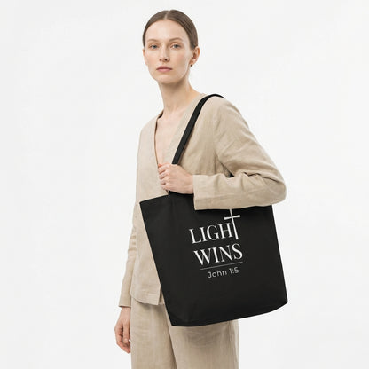 Lifestyle photo of a model holding a black tote bag featuring “Light Wins,” a cross graphic, and “John 1:5.”