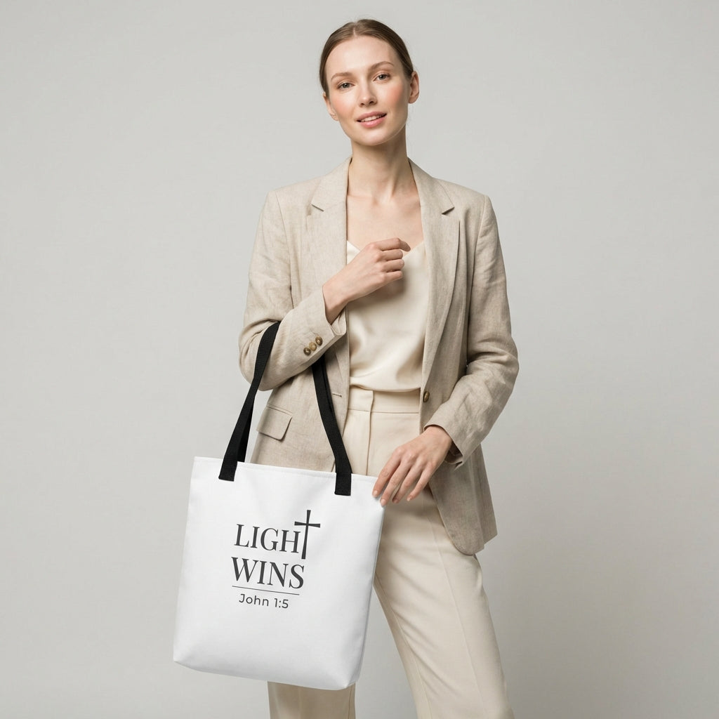 Model holding a white tote bag with dark gray “Light Wins” and cross design, John 1:5.