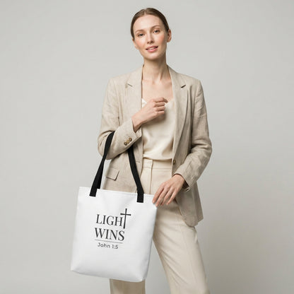 Model holding a white tote bag with dark gray “Light Wins” and cross design, John 1:5.