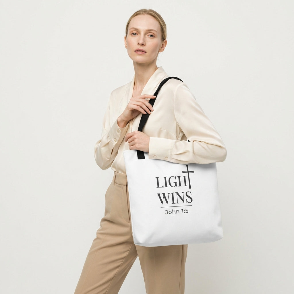 Model holding a white tote bag with dark gray “Light Wins” and cross design, John 1:5.