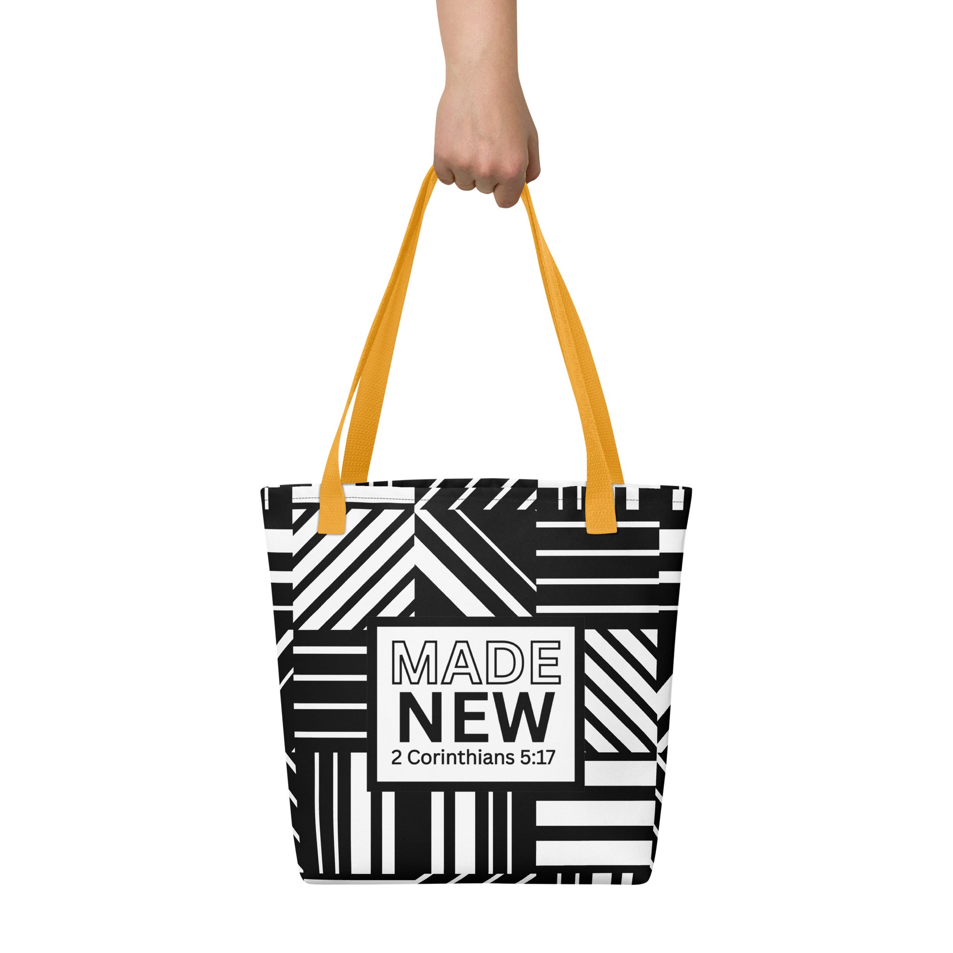 Tote bag carried by hand with yellow straps, featuring the bold black-and-white pattern and the “MADE NEW — 2 Corinthians 5:17” label.
