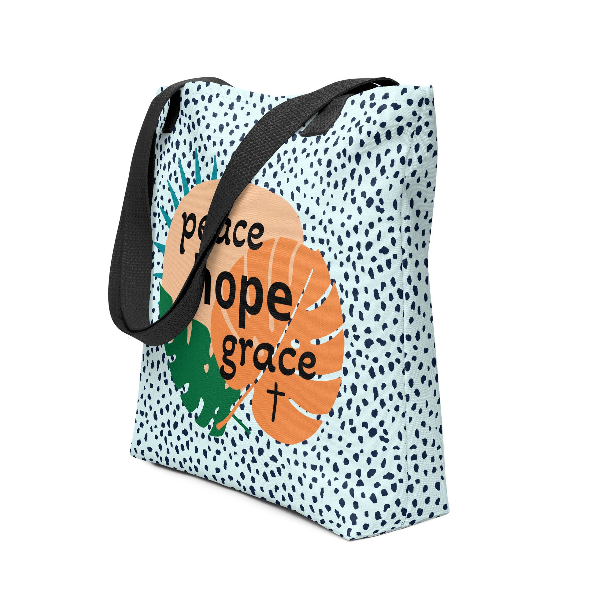 Angled view of tote bag with black handles, light blue speckle print, and “peace hope grace” graphic over tropical leaves.