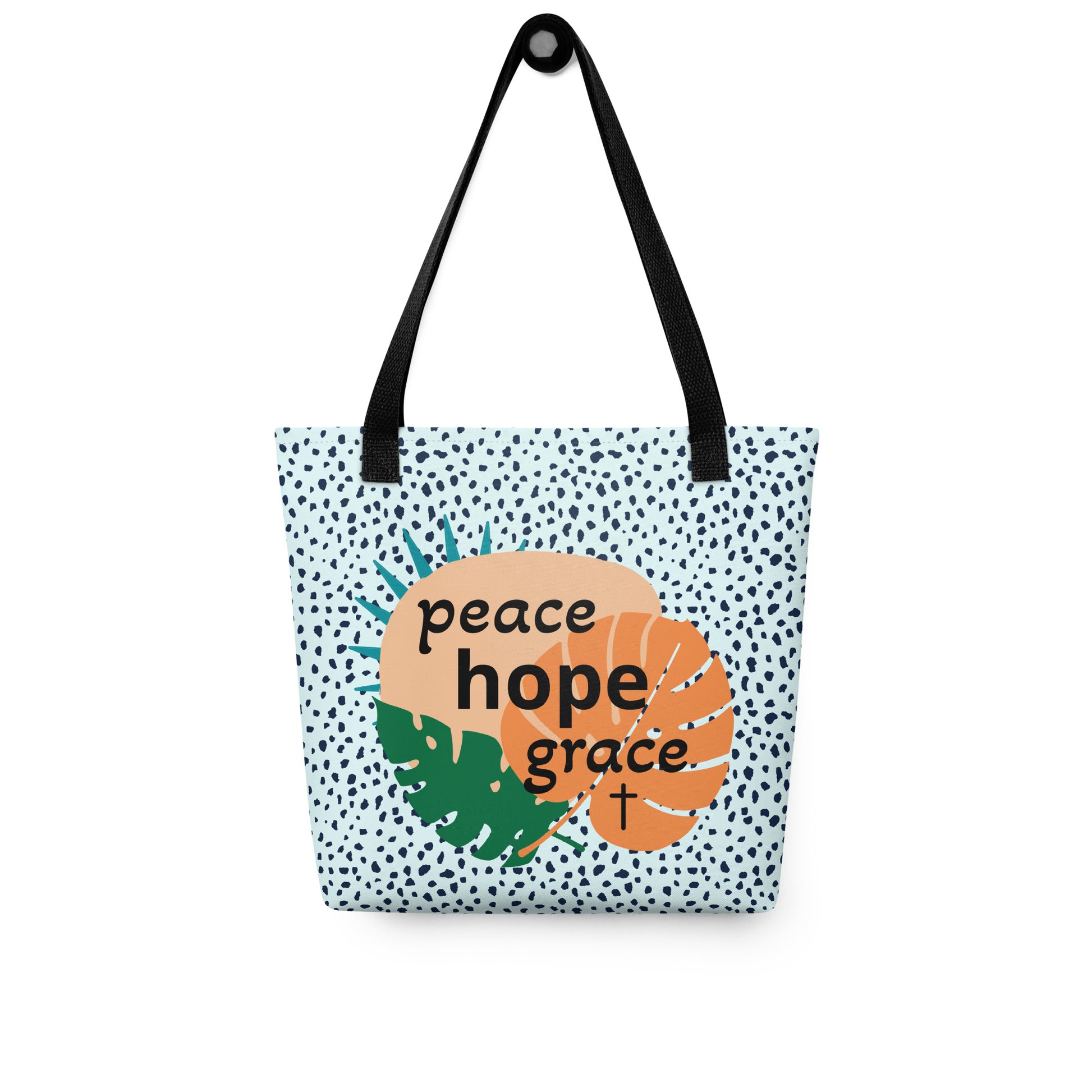 Tote bag hanging on a hook with black straps, blue speckled fabric, and “peace hope grace” tropical leaf design with a cross.