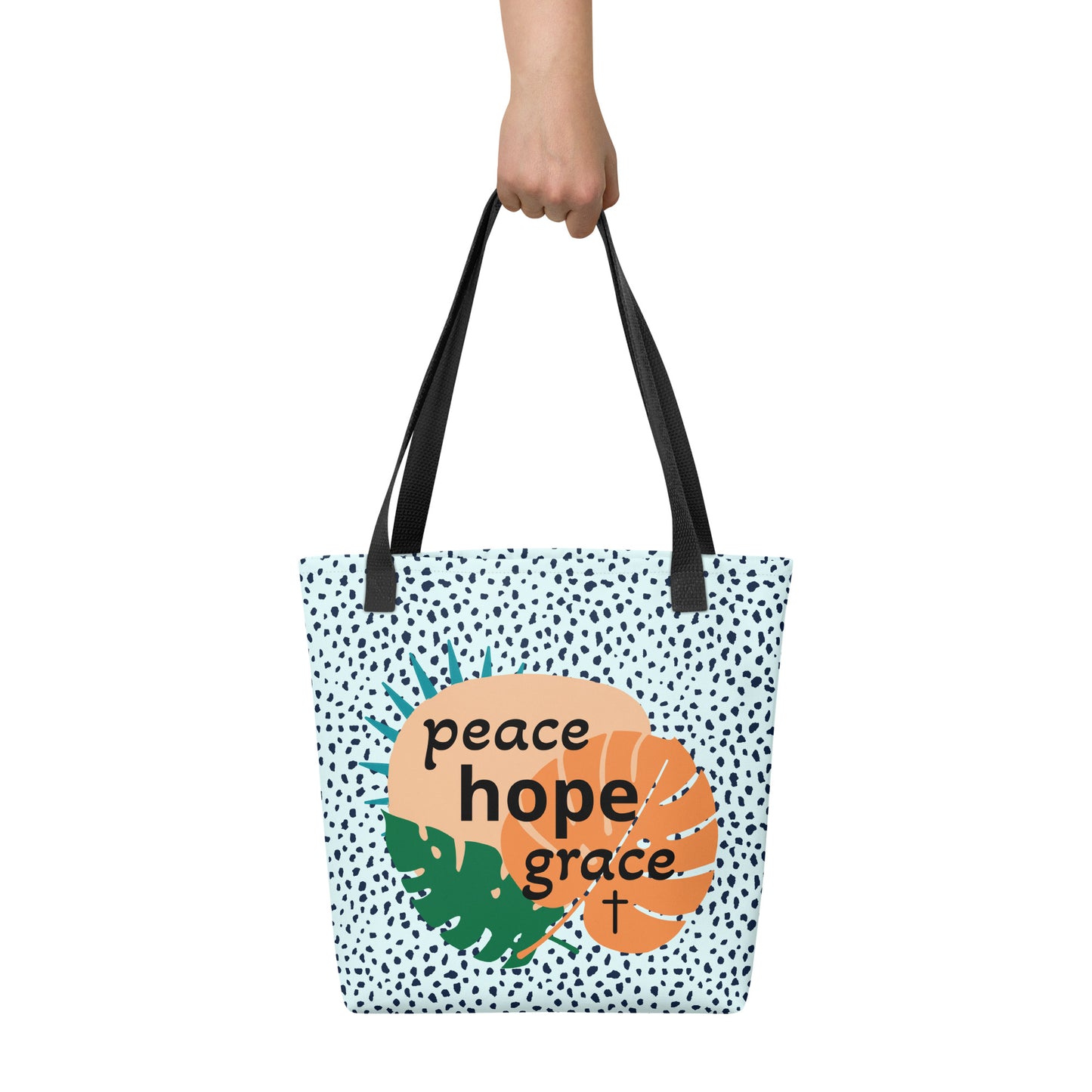 Hand holding tote bag with black handles, light blue speckle pattern, and “peace hope grace” graphic with tropical leaves and a cross.