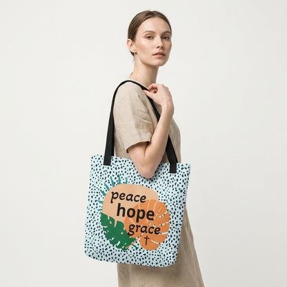 Lifestyle photo of a person carrying the tote bag with black straps, blue speckle pattern, and “peace hope grace” tropical leaf graphic with a cross.