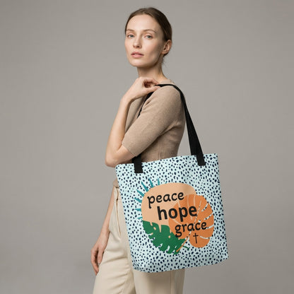 Lifestyle photo of a person carrying the tote bag with black straps, blue speckle pattern, and “peace hope grace” tropical leaf graphic with a cross.