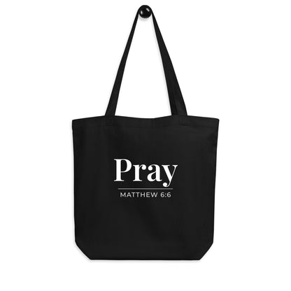 Black cotton tote bag hanging on a hook featuring “Pray” and “Matthew 6:6” in white text.