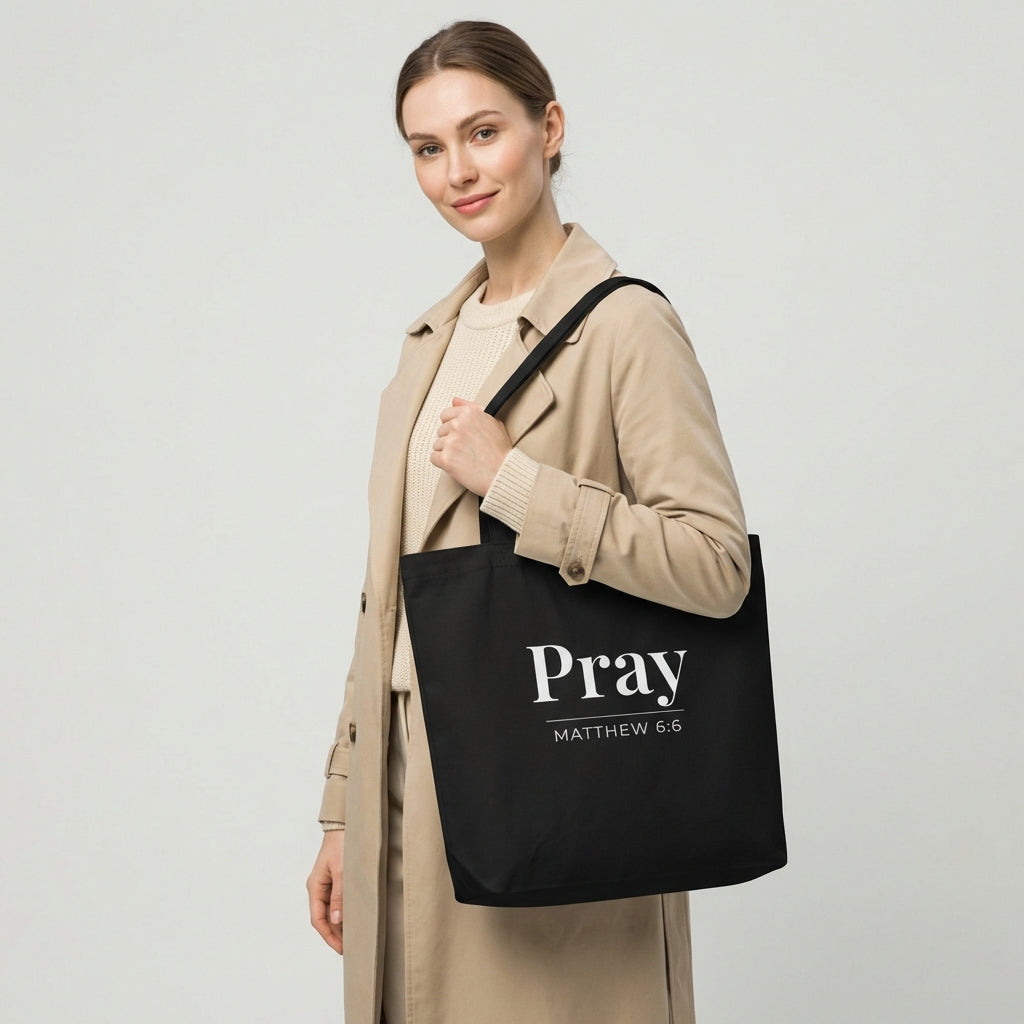 Model holding a black cotton tote bag printed with “Pray” and “Matthew 6:6” in white text.