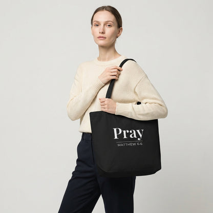 Model holding a black cotton tote bag printed with “Pray” and “Matthew 6:6” in white text.