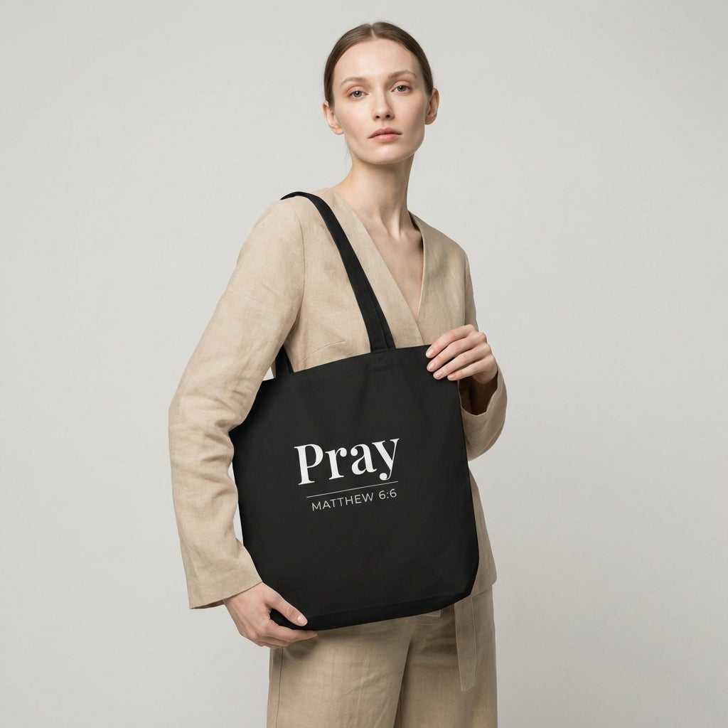 Model holding a black cotton tote bag printed with “Pray” and “Matthew 6:6” in white text.