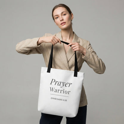 Lifestyle photo of a model holding a white tote bag with black handles featuring “Prayer Warrior” and “Ephesians 6:18” in dark gray.