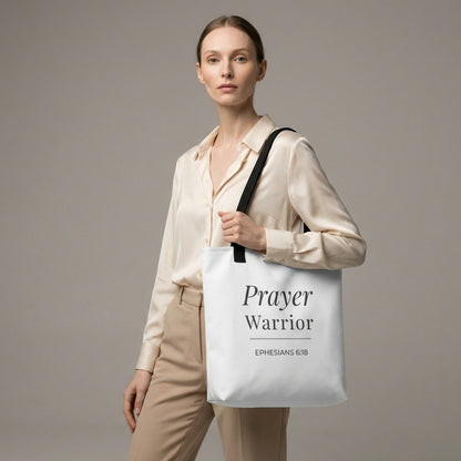 Lifestyle photo of a model holding a white tote bag with black handles featuring “Prayer Warrior” and “Ephesians 6:18” in dark gray.