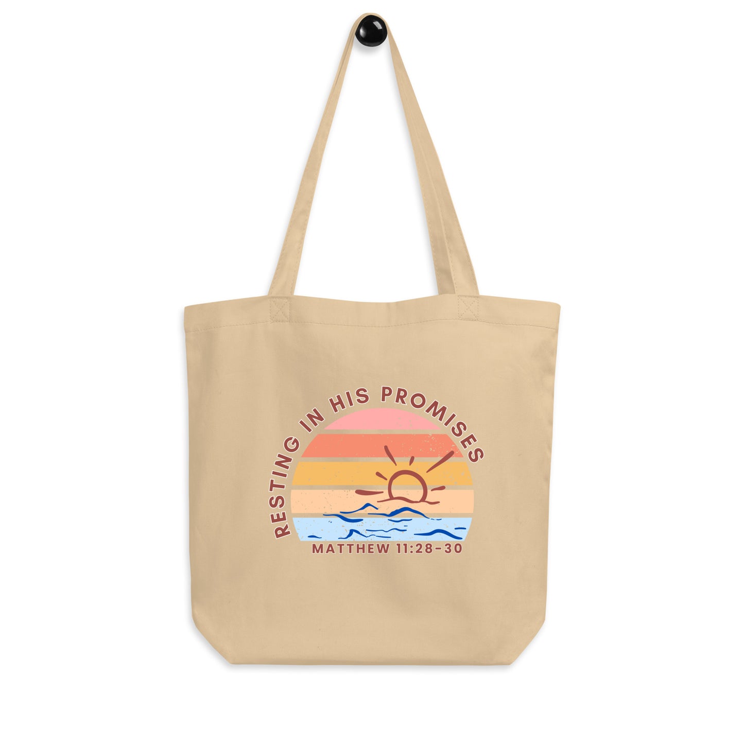A cotton tote bag hanging on a hook featuring “Resting in His Promises,” “Matthew 11:28–30,” and a sunset-over-waves graphic.