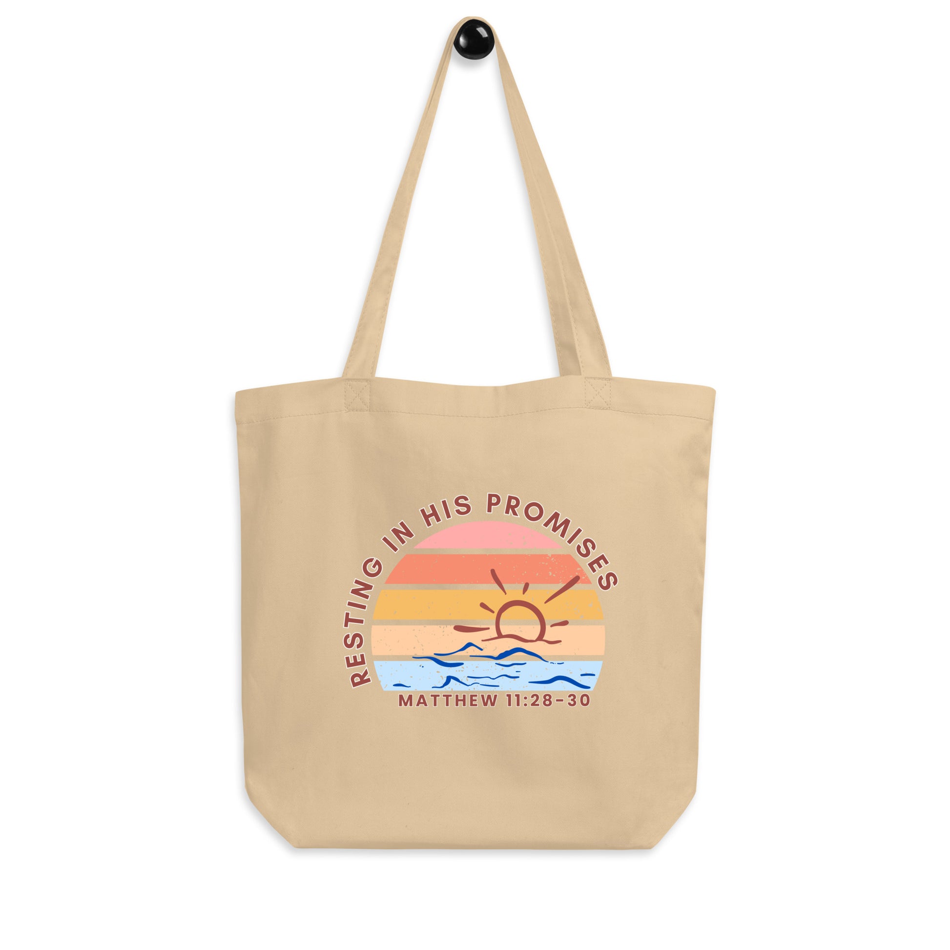 A cotton tote bag hanging on a hook featuring “Resting in His Promises,” “Matthew 11:28–30,” and a sunset-over-waves graphic.