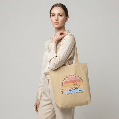 Lifestyle photo of a model holding a cotton tote bag featuring “Resting in His Promises” and “Matthew 11:28–30” with a sunset wave design.