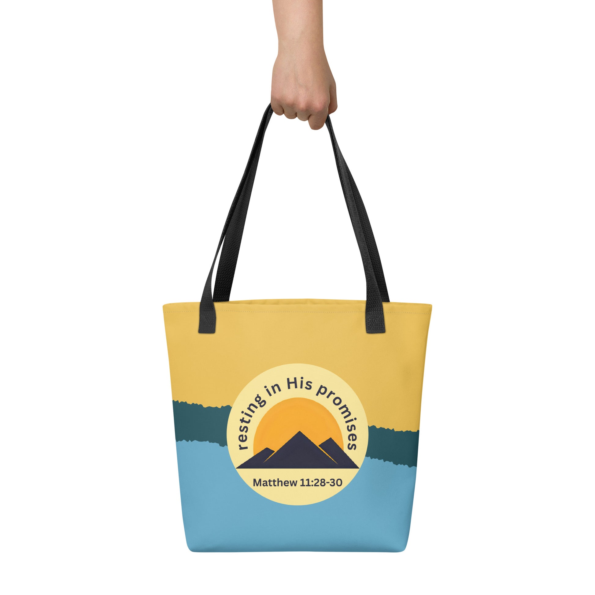 Hand holding tote bag with black handles, yellow and blue color block, and “resting in His promises” mountain graphic with Matthew 11:28–30.