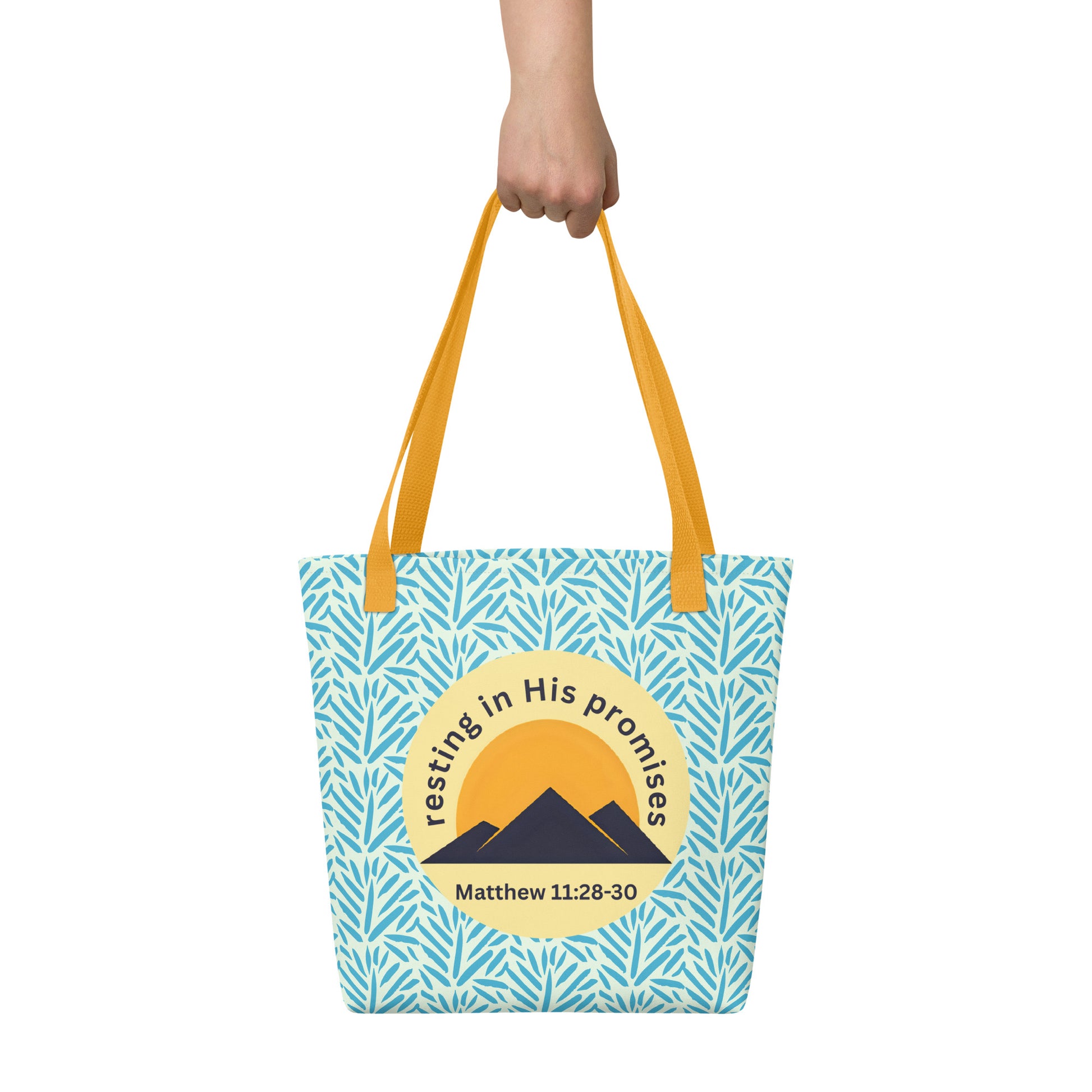 Hand holding “Resting in His Promises” tote bag with yellow straps, blue leaf print background, and Matthew 11:28–30 mountain badge.