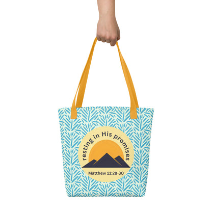 Hand holding “Resting in His Promises” tote bag with yellow straps, blue leaf print background, and Matthew 11:28–30 mountain badge.