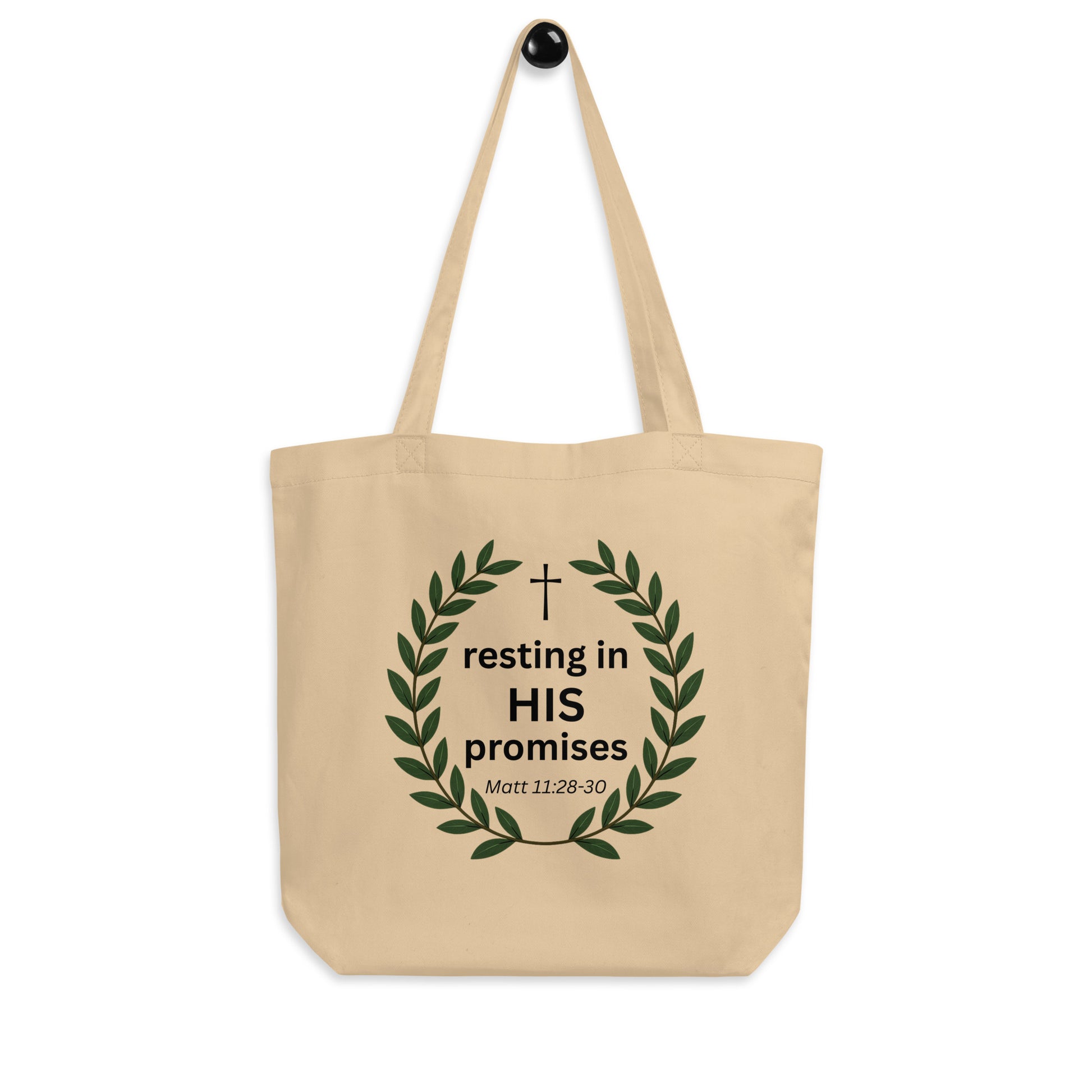 Natural cotton tote bag hanging on a hook with a leafy wreath design and “resting in HIS promises” (Matt 11:28–30) printed on the front.