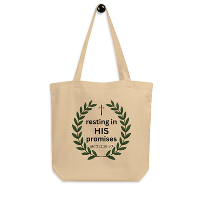 Natural cotton tote bag hanging on a hook with a leafy wreath design and “resting in HIS promises” (Matt 11:28–30) printed on the front.