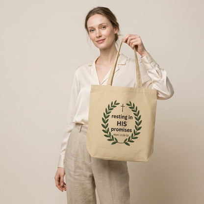 Model holding a natural cotton tote bag featuring a leafy wreath, cross, and “resting in HIS promises” (Matt 11:28–30) design.