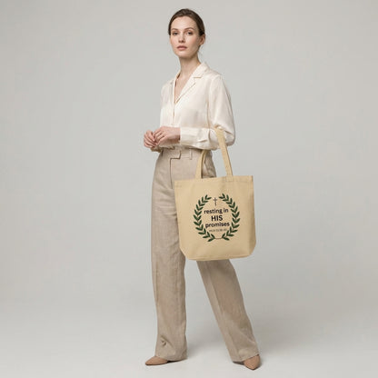 Model holding a natural cotton tote bag featuring a leafy wreath, cross, and “resting in HIS promises” (Matt 11:28–30) design.