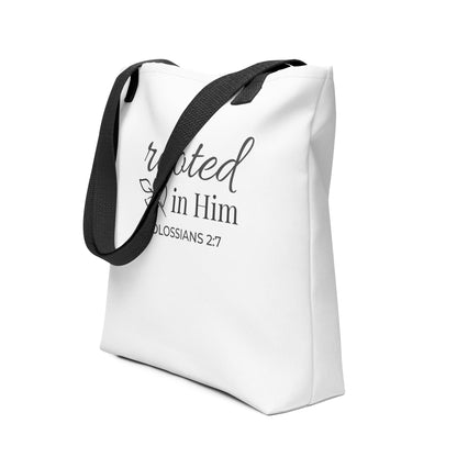 Angled view of a white tote bag with black handles featuring “rooted in Him” and “Colossians 2:7” in dark gray with a leaf design.