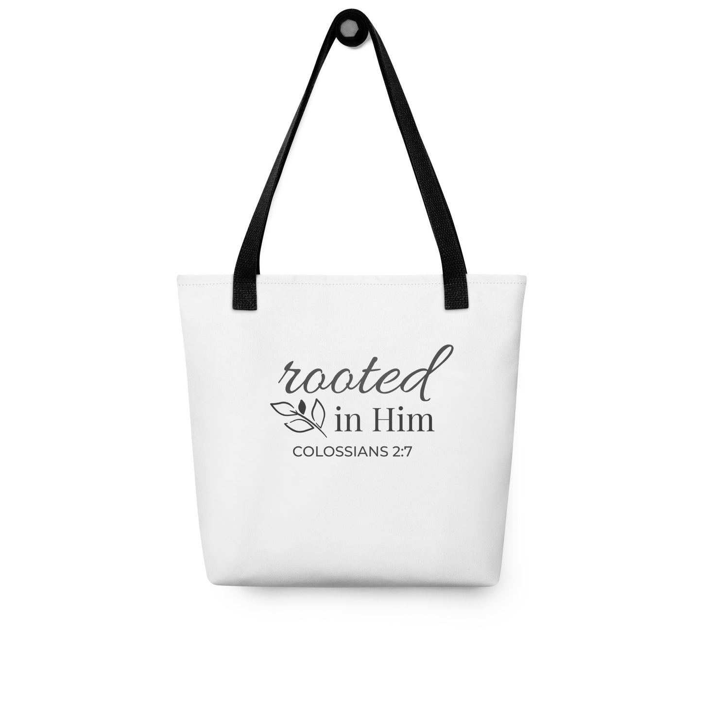 White tote bag hanging on a hook with black handles featuring “rooted in Him” and “Colossians 2:7” in dark gray with a leaf design.