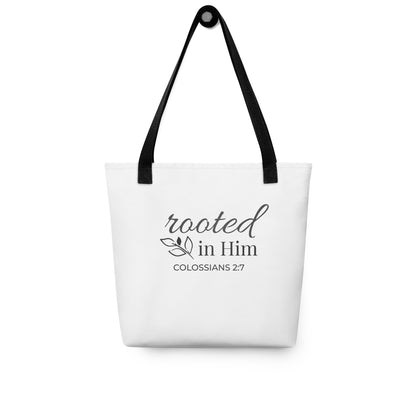 White tote bag hanging on a hook with black handles featuring “rooted in Him” and “Colossians 2:7” in dark gray with a leaf design.