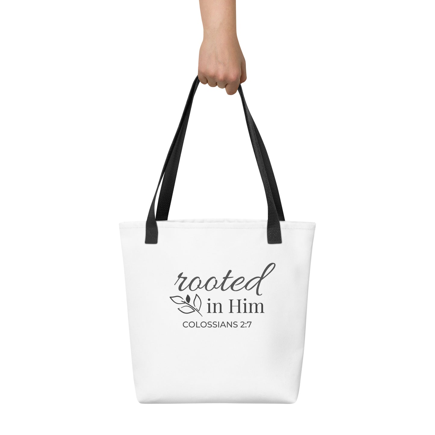 White tote bag held in hand with black handles featuring “rooted in Him” and “Colossians 2:7” in dark gray with a leaf design.