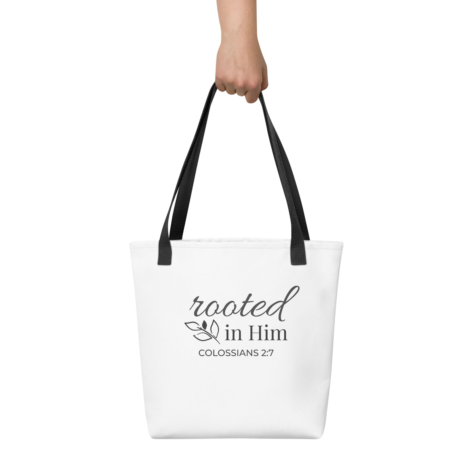 White tote bag held in hand with black handles featuring “rooted in Him” and “Colossians 2:7” in dark gray with a leaf design.