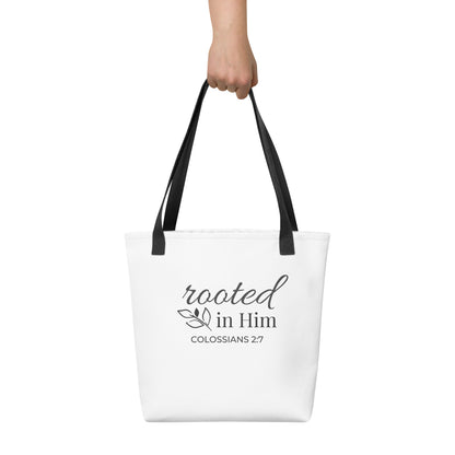 White tote bag held in hand with black handles featuring “rooted in Him” and “Colossians 2:7” in dark gray with a leaf design.