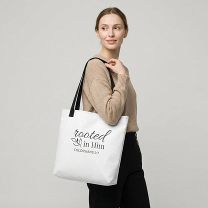 Lifestyle photo of a model holding a white tote bag with black handles featuring “rooted in Him” and “Colossians 2:7” with a leaf design.