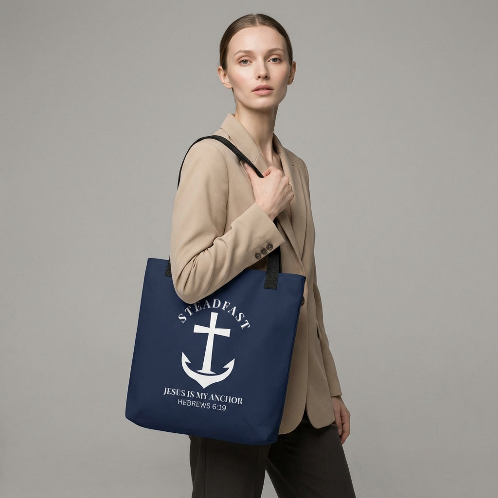 Model carrying a navy tote with black straps, featuring a white anchor cross graphic and the message “STEADFAST — JESUS IS MY ANCHOR” (Hebrews 6:19) on the front.